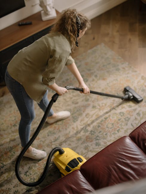 A woman with curly hair wearing a green jacket and jeans is using a handheld vacuum cleaner to deep clean an ornate, patterned carpet in a living room. The room features wooden flooring, a brown leath
