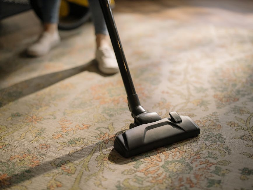 A woman with curly hair wearing a green jacket and jeans is using a handheld vacuum cleaner to deep clean an ornate, patterned carpet in a living room. The room features wooden flooring, a brown leath